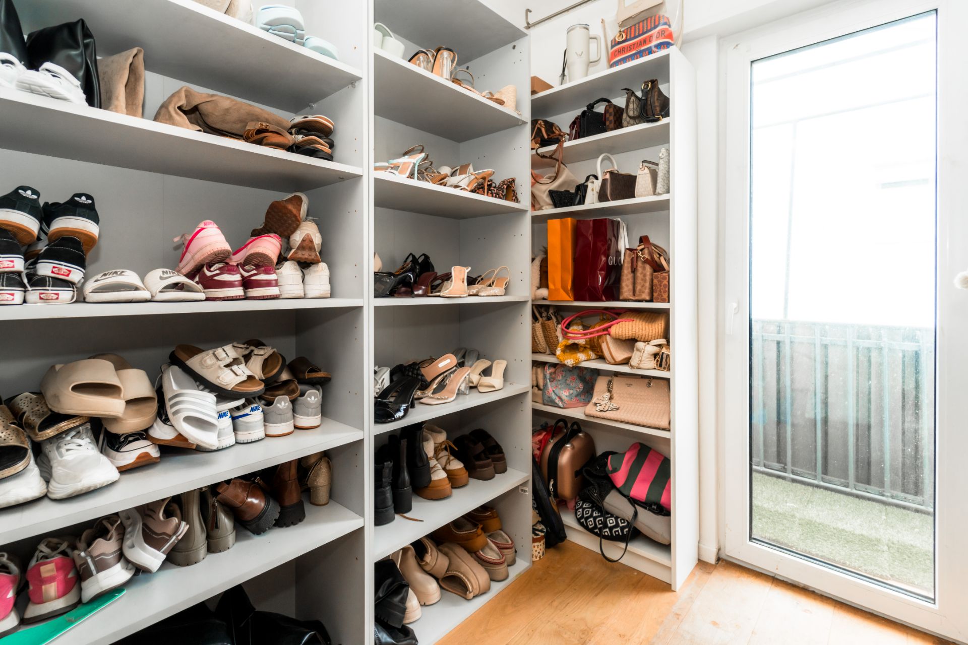 Ein Raum mit weißen Regalen voller Schuhe, Taschen und Handtaschen neben einer Glastür.