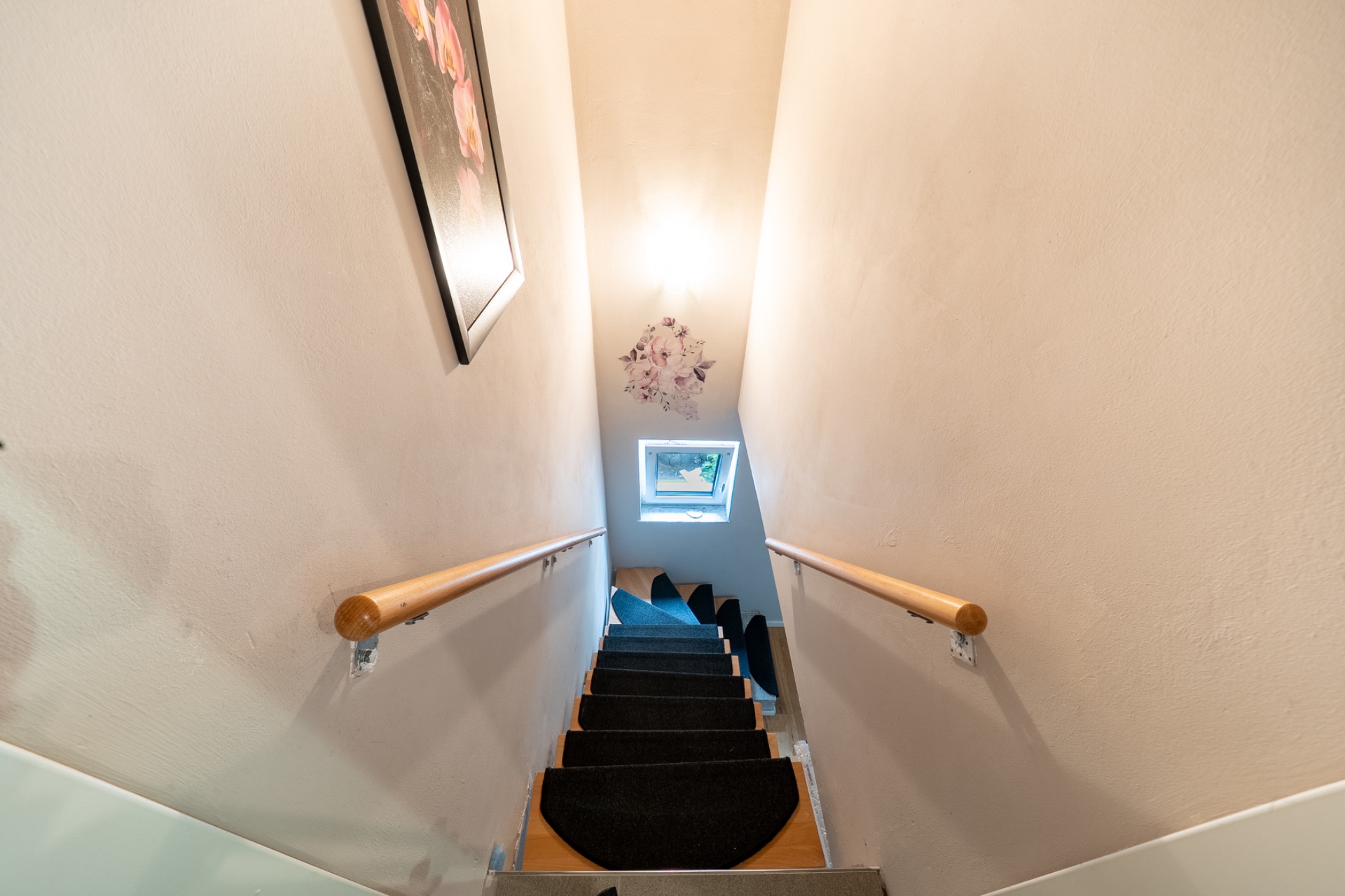 Schmale Treppe mit Handläufen, kleinem Fenster am Ende und Bild an der Wand.