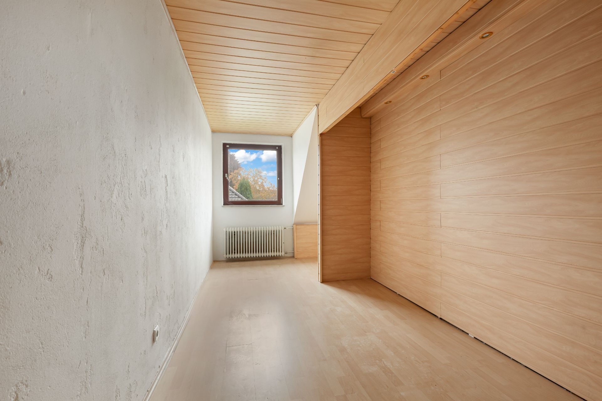Schmales, leeres Zimmer mit Holzdecke, Holzwand und Fenster am Ende.