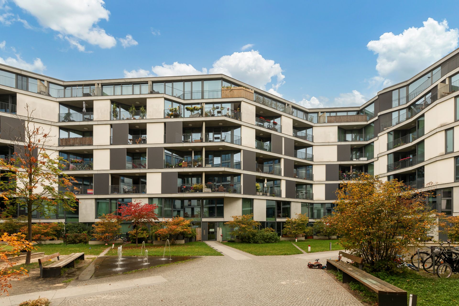 Modernes Mehrfamilienhaus mit Balkonen und Innenhof, umgeben von Bäumen und Sitzbänken.