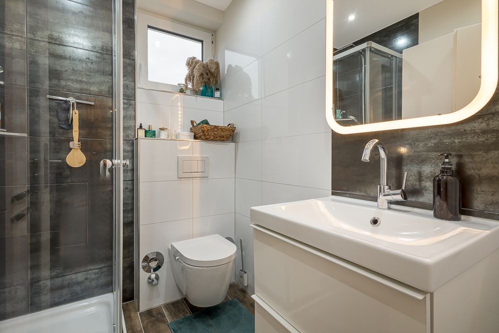 Modernes Badezimmer mit Dusche, WC, Waschbecken, beleuchtetem Spiegel und Regal.