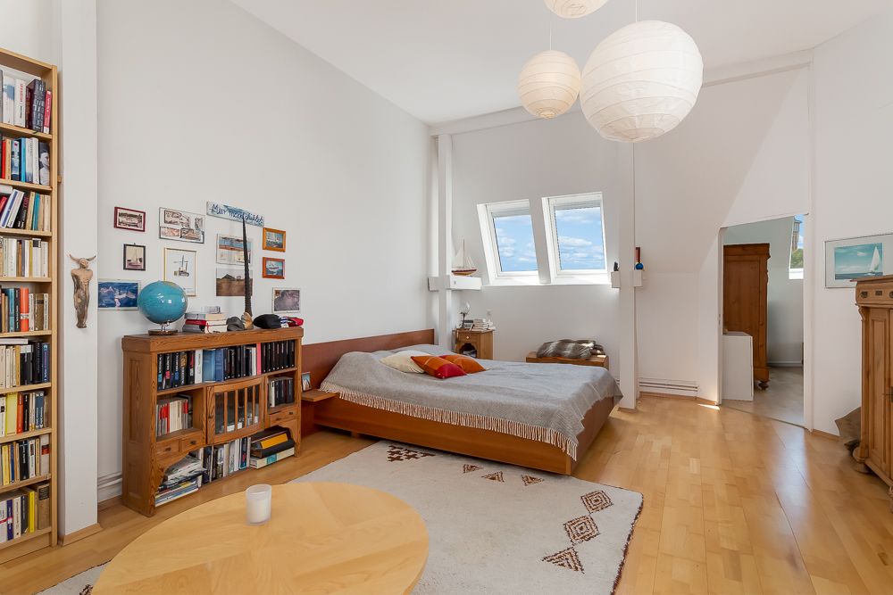 Helles Schlafzimmer mit Holzboden, Bücherregal, Bett, Bildern an der Wand und zwei Dachfenstern.