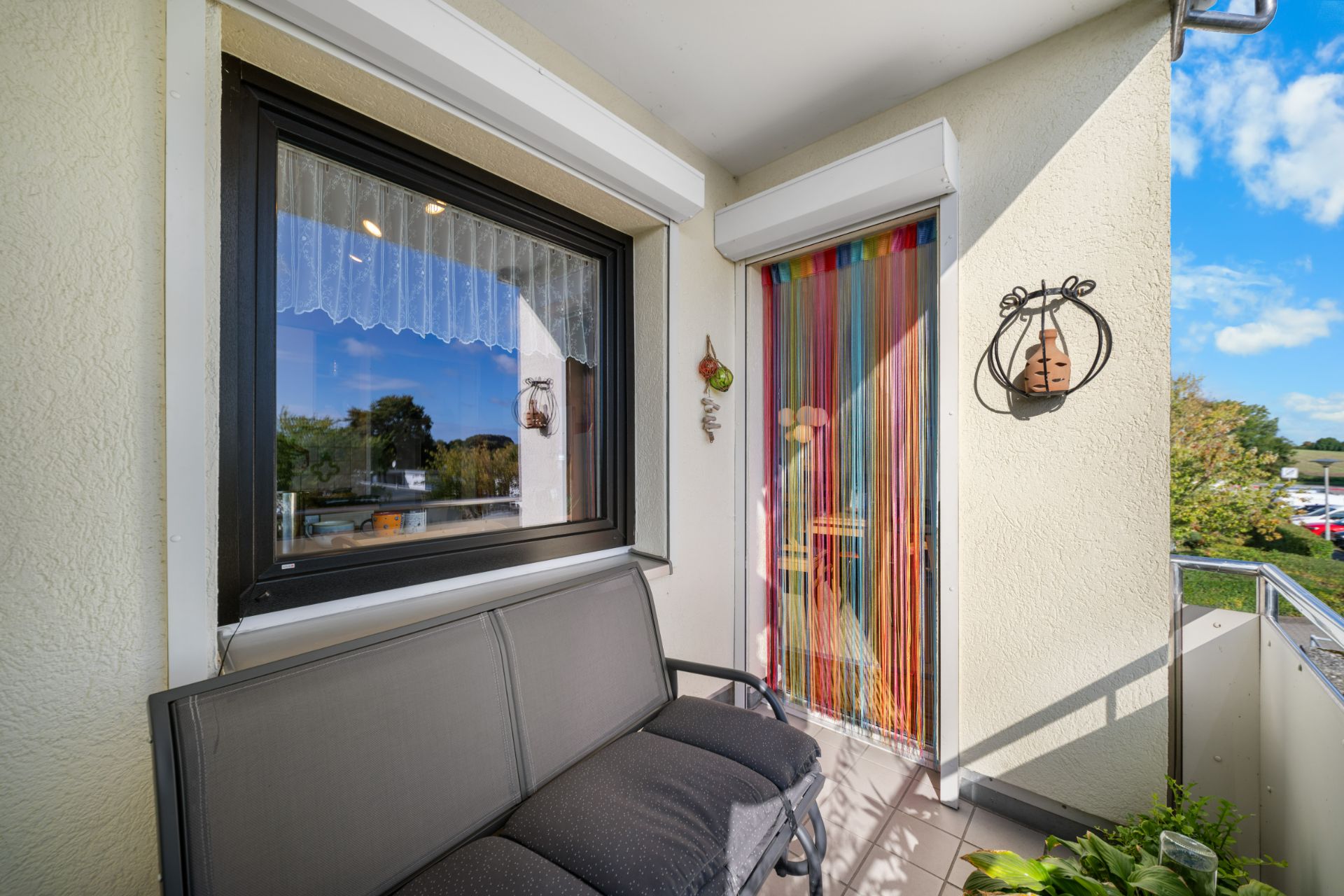 Kleiner Balkon mit grauer Bank, Fenster, bunter Perlenschnur und Wanddeko.