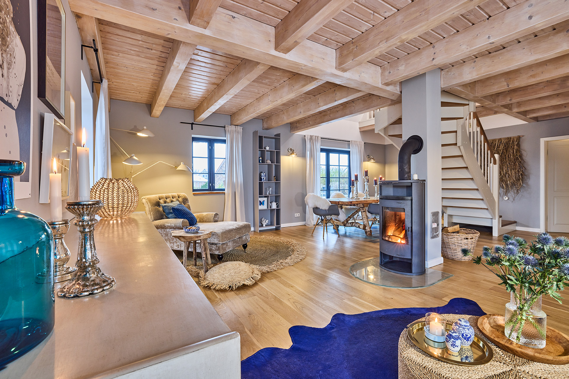 Gemütliches Wohnzimmer mit Holzdecke, Kamin, Essbereich und Treppe, viele Dekorationen und große Fenster.