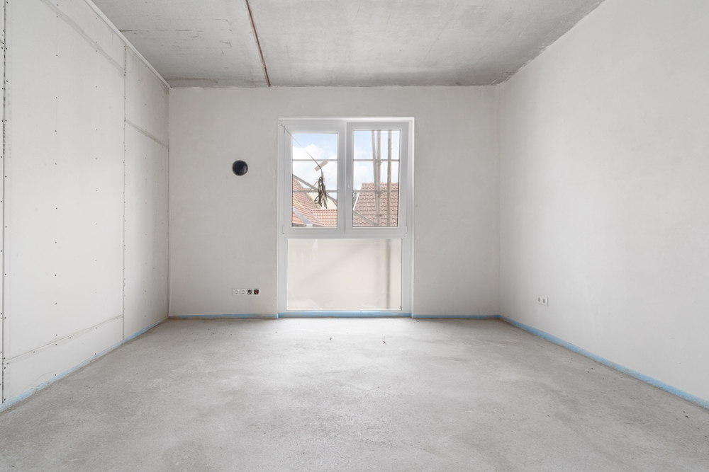 Leerer, unfertiger Raum mit weiß gestrichenen Wänden, Fenster und sichtbarem Betonboden.