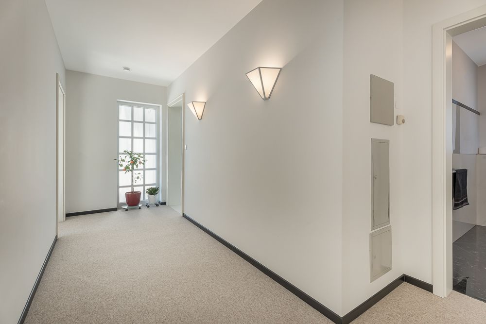 Heller Flur mit Teppichboden, Wandlampen und einem Fenster mit Topfpflanzen am Ende.