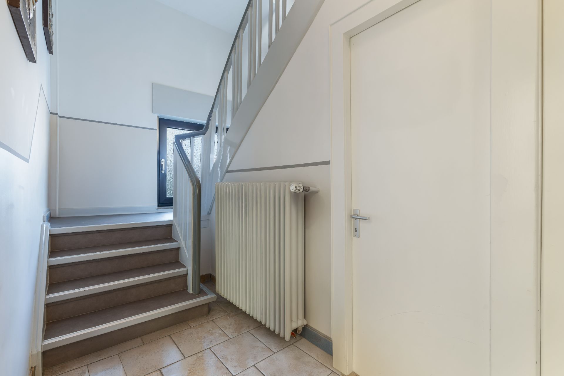 Heller Hausflur mit Treppe, Heizung, Fliesenboden und einer weißen Tür.