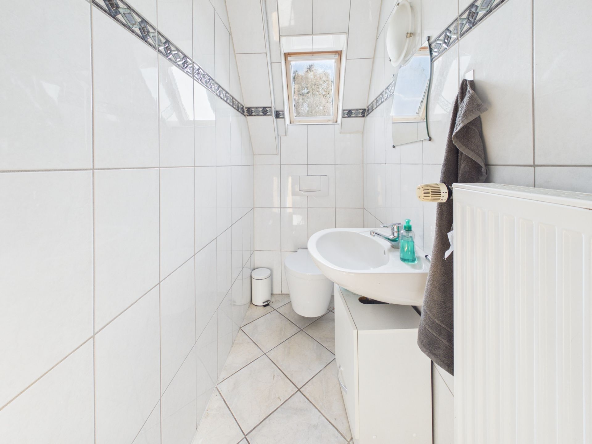 Helles, gefliestes Bad mit Waschbecken, Toilette, Fenster und Heizkörper.