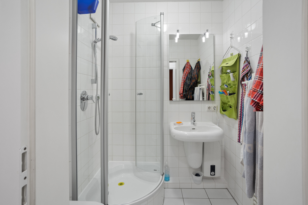 Kleines Badezimmer mit Dusche, Waschbecken, Spiegel und bunten Handtüchern an der Wand.
