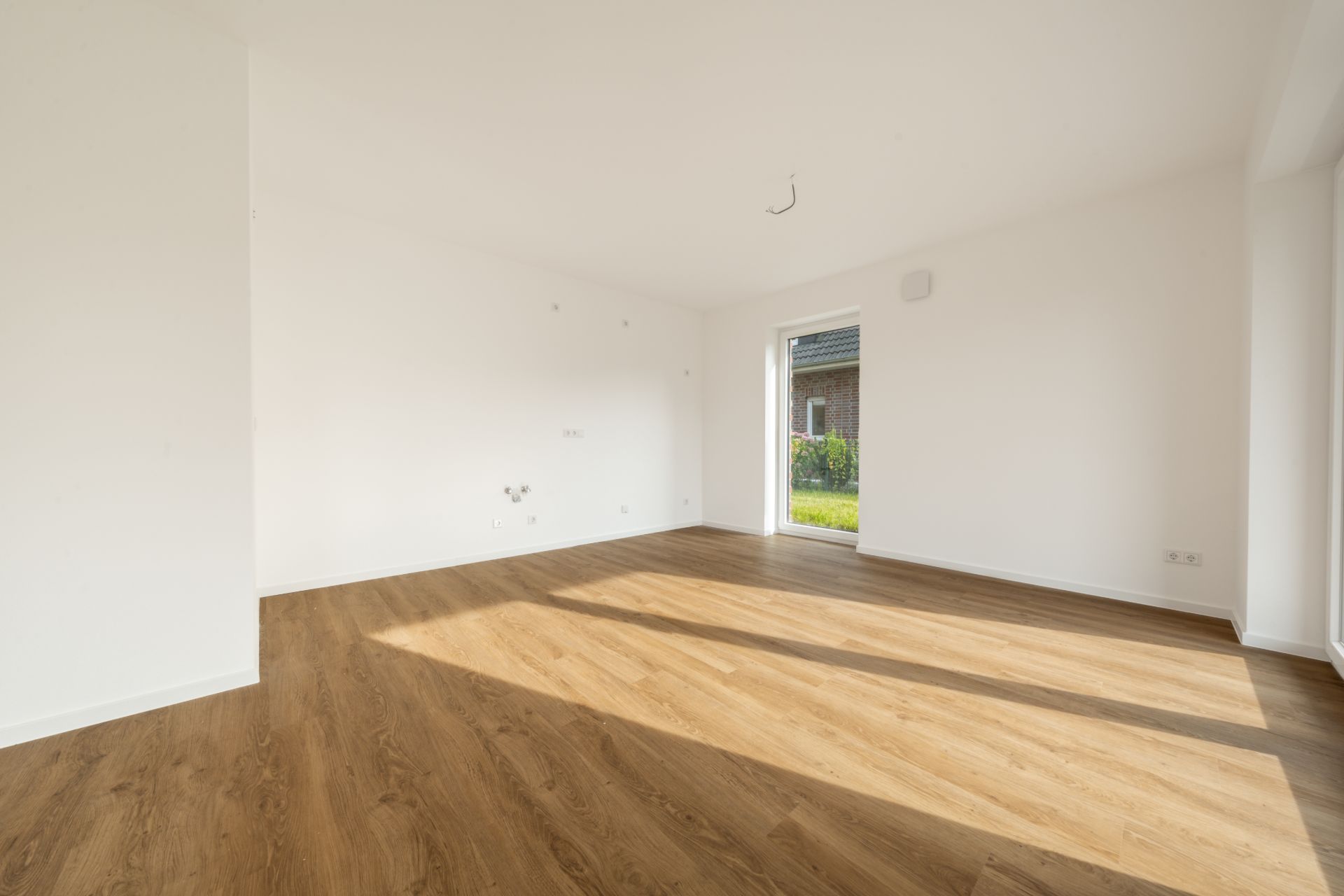 Heller leerer Raum mit Holzfußboden und weißen Wänden, Tür führt nach draußen.