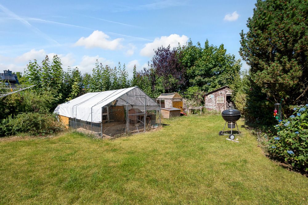 Garten mit Gewächshaus, kleinen Holzhütten und einem Grill auf einer Rasenfläche.