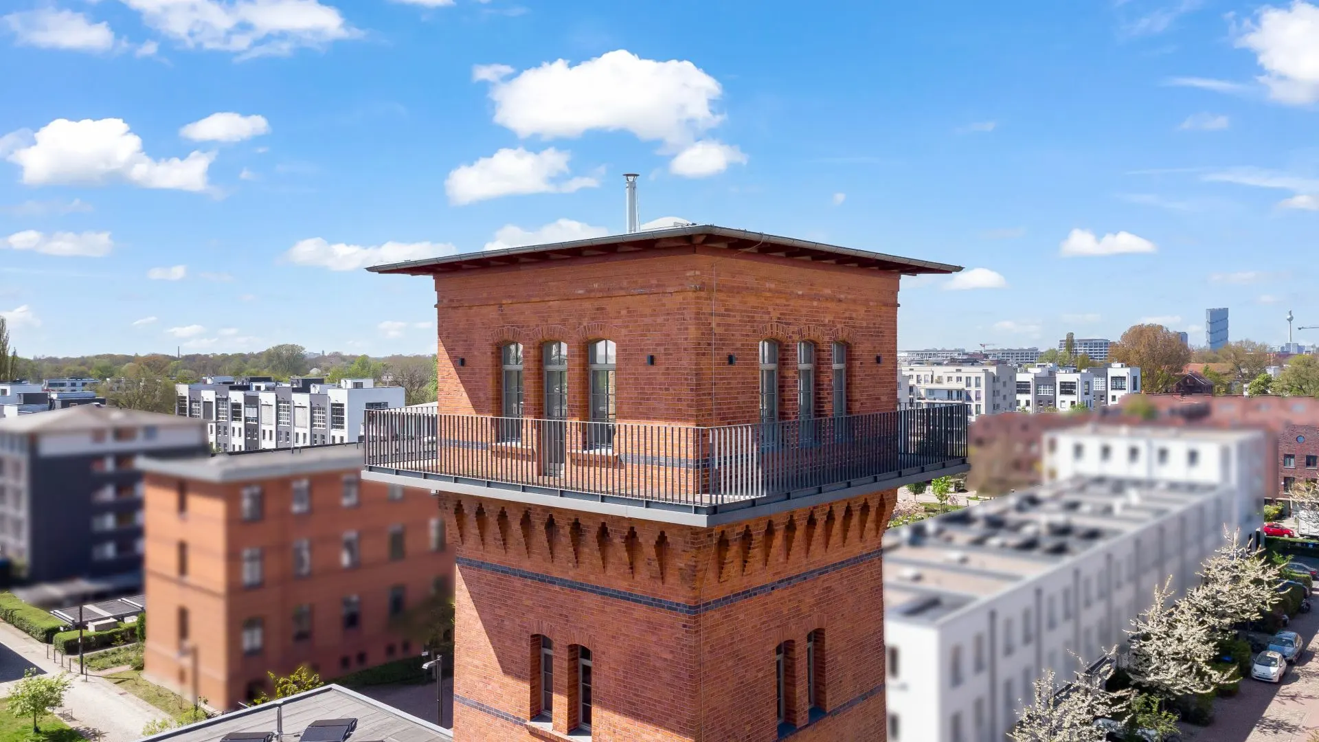 Backsteinturm mit umlaufendem Balkon, umgeben von modernen Wohngebäuden bei blauem Himmel.