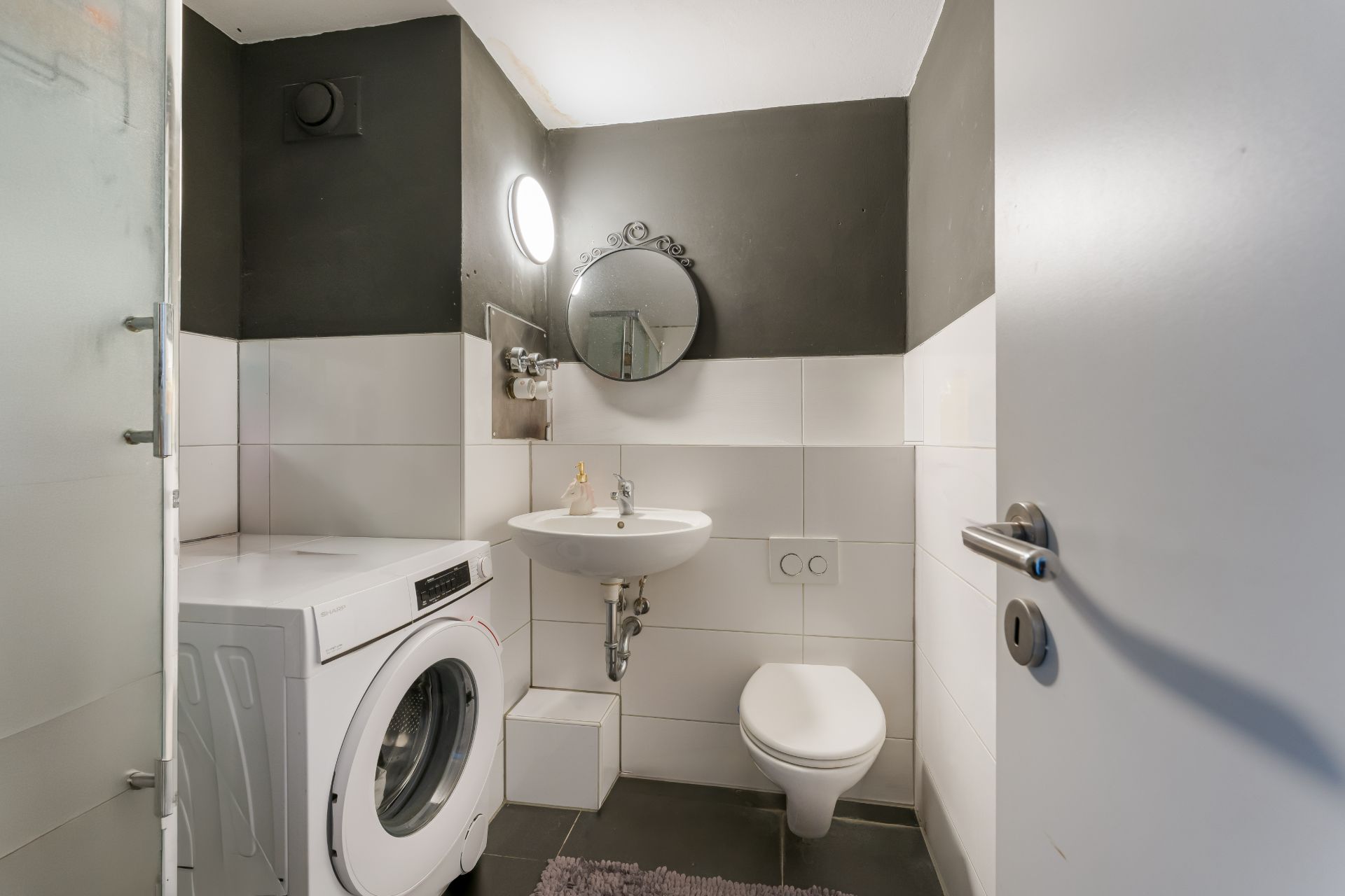 Kleines Badezimmer mit Waschmaschine, Waschbecken, Spiegel und Wandtoilette.