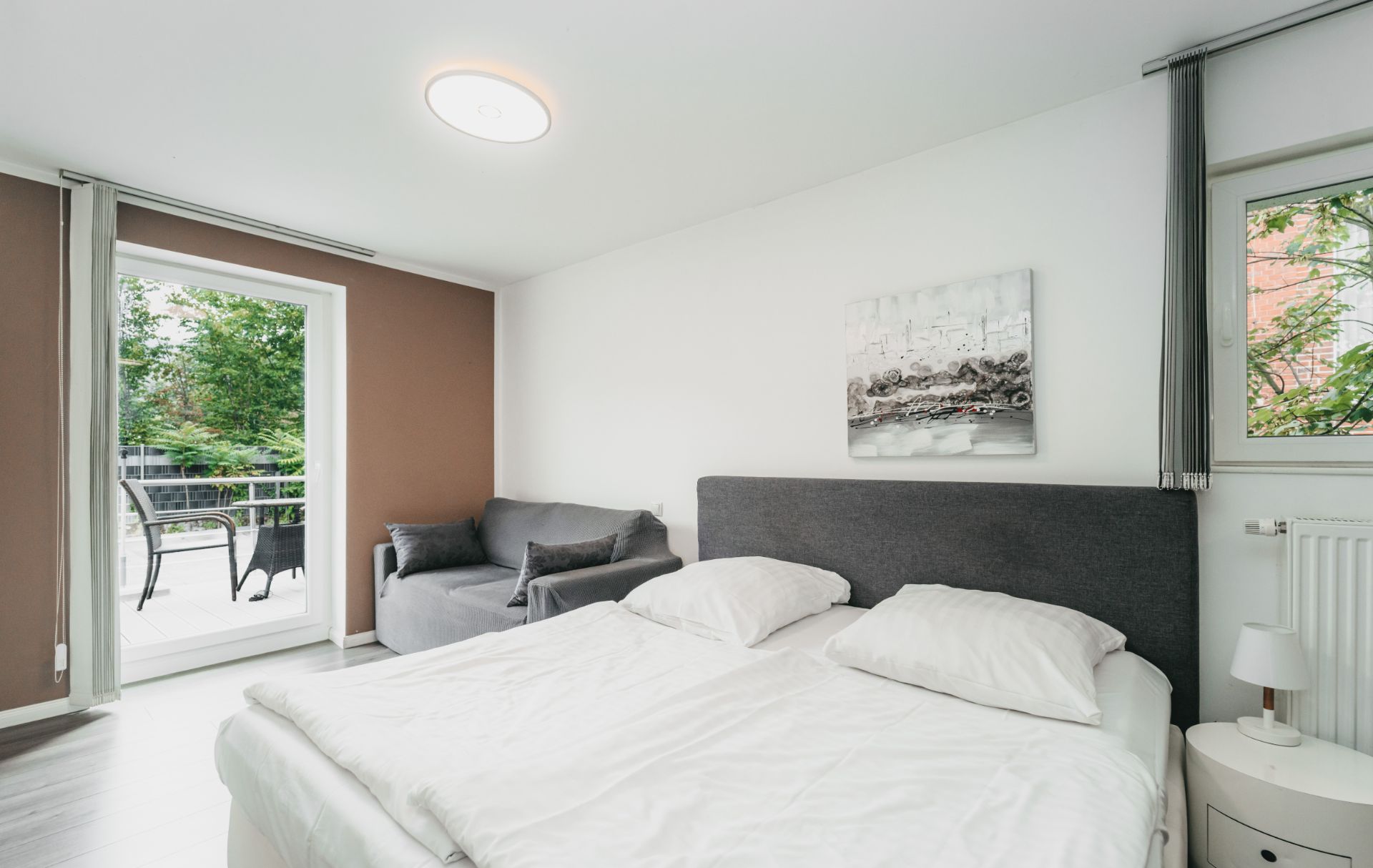 Helles Schlafzimmer mit Doppelbett, grauem Sofa und Zugang zu einem Balkon mit Blick ins Grüne.