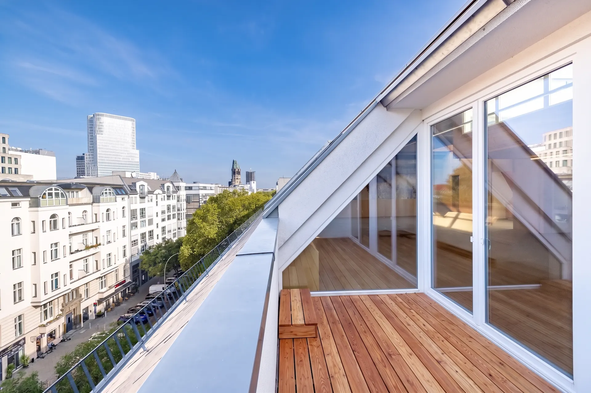 Dachterrasse mit Holzbelag und Glasfassade, Blick auf Stadtgebäude und blauen Himmel.