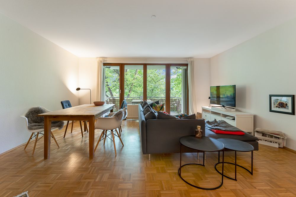 Helles Wohnzimmer mit Sofa, Esstisch, Fernseher und großen Fenstern mit Blick ins Grüne.