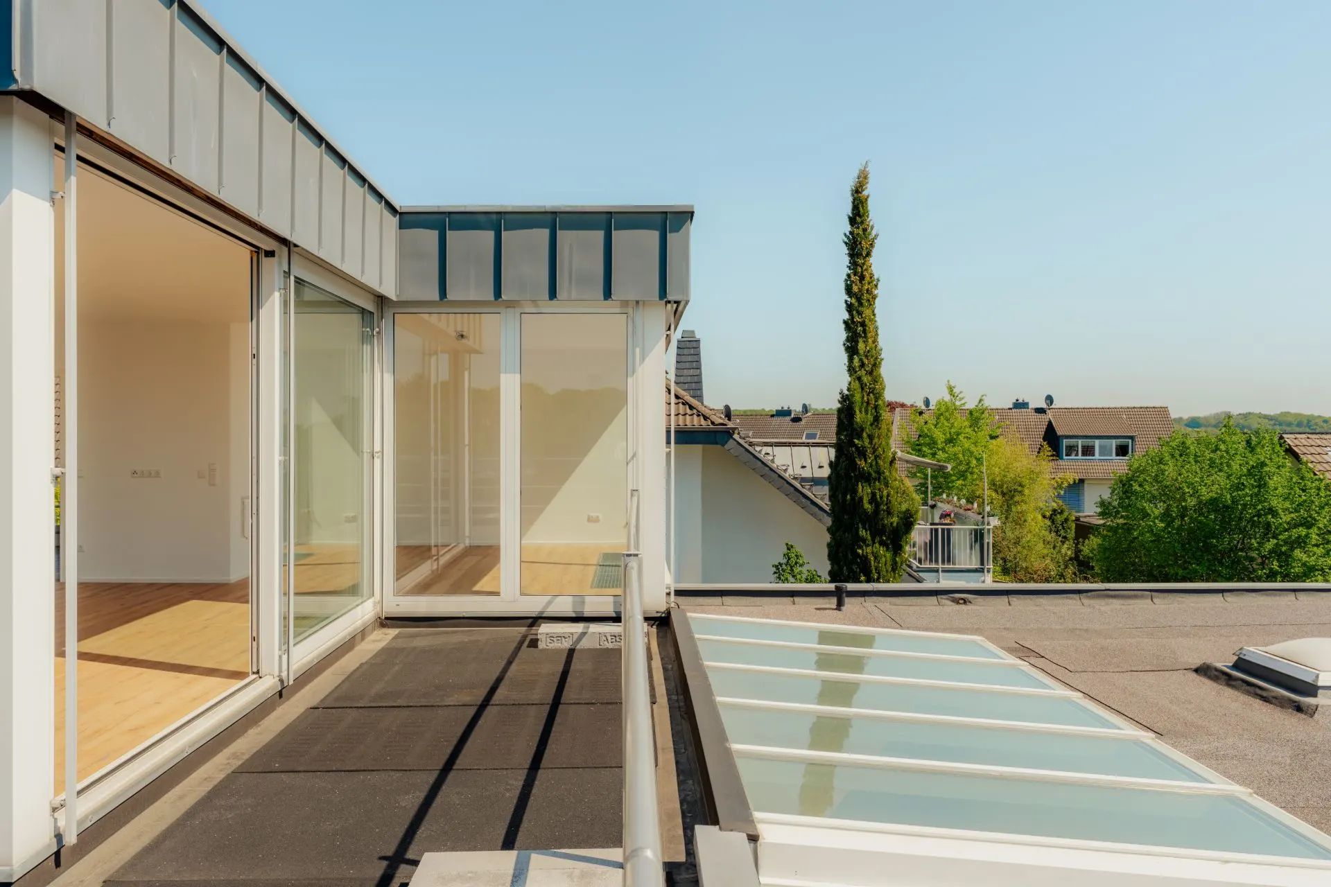 Dachterrasse mit Glasgeländer und großen Fenstern, Blick auf Nachbarhäuser und Bäume.