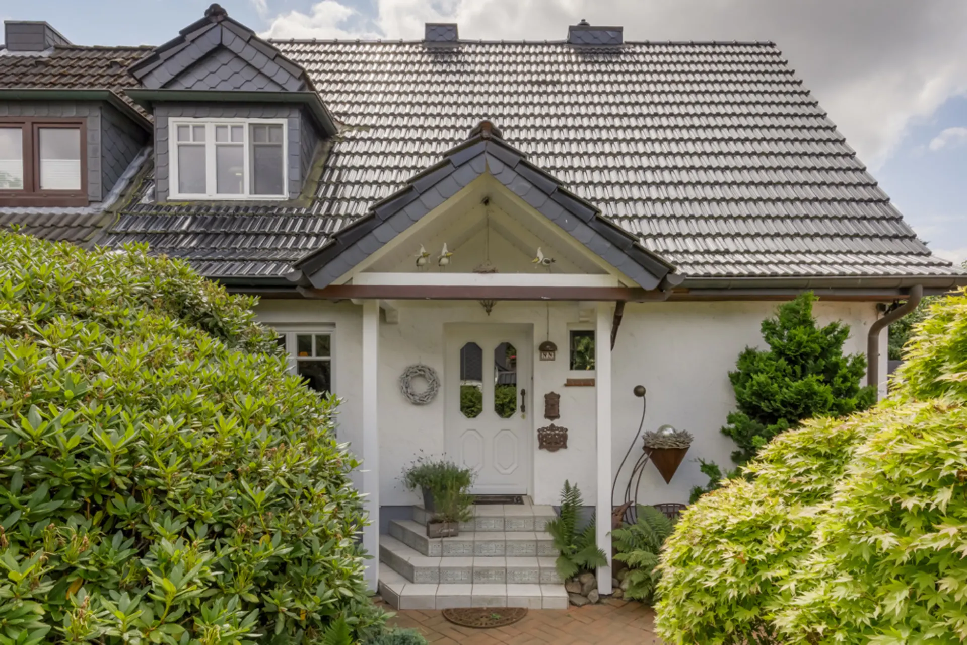 Eingang eines Hauses mit weißer Tür, Stufen und umgebenden grünen Büschen.
