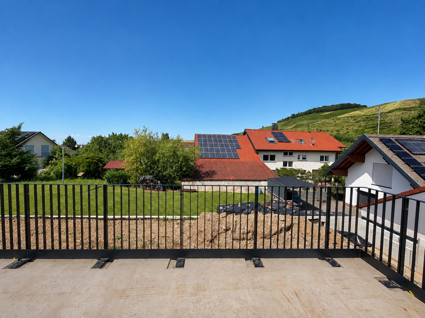 Blick von einer Terrasse mit Geländer auf Gärten und Häuser mit Solarpaneelen.