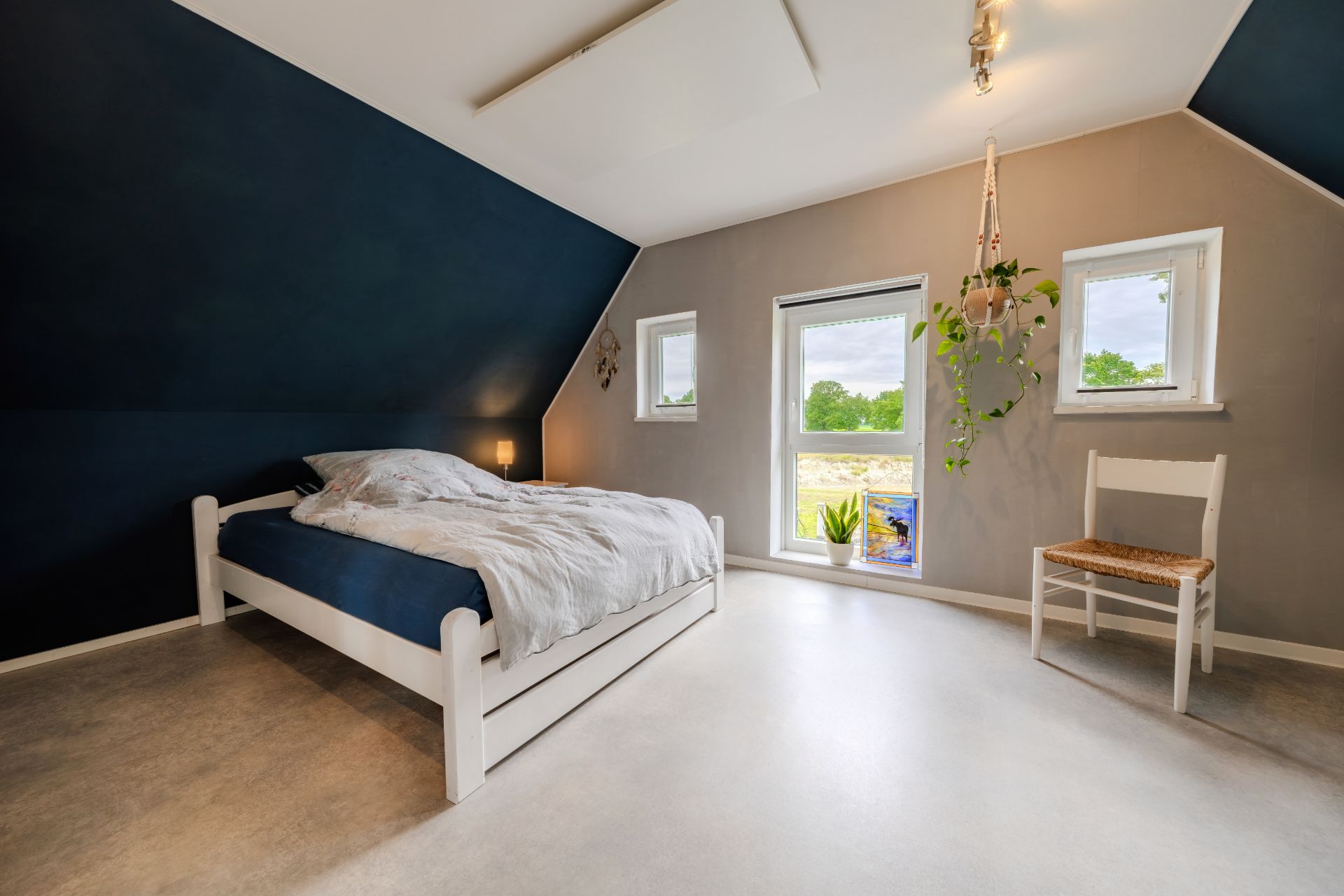 Schlafzimmer mit blau-weißen Wänden, Bett, Stuhl und drei Fenstern mit Blick ins Grüne.