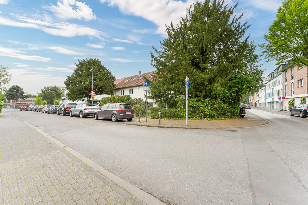 Straßenecke mit parkenden Autos, Wohnhäusern und großen Bäumen bei Tageslicht.
