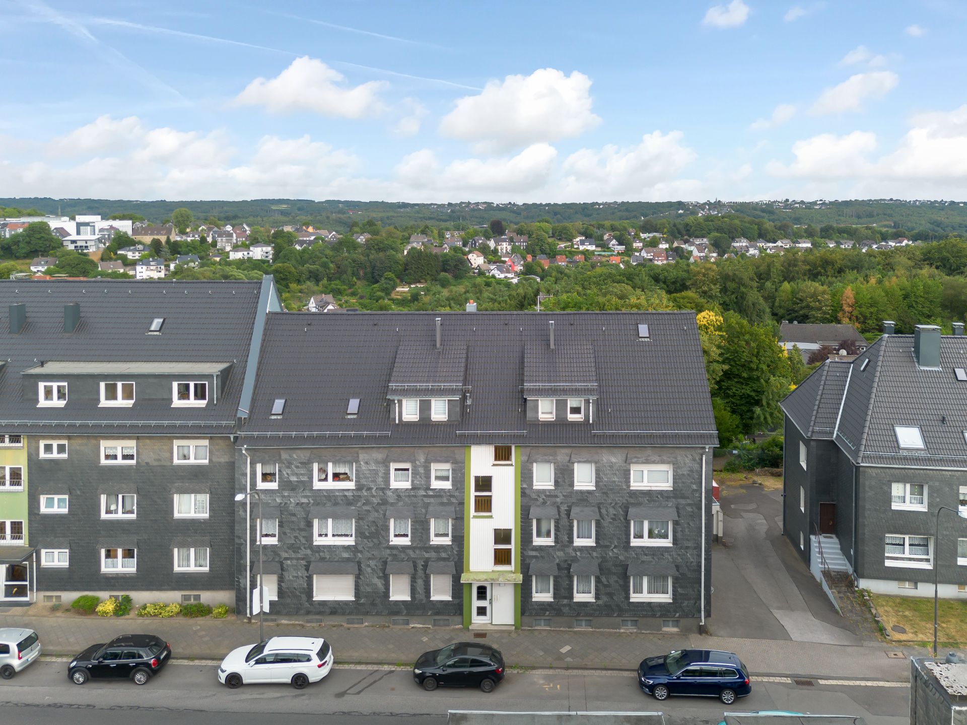 Mehrfamilienhaus mit mehrere Stockwerken und geparkten Autos davor, Bäume im Hintergrund.
