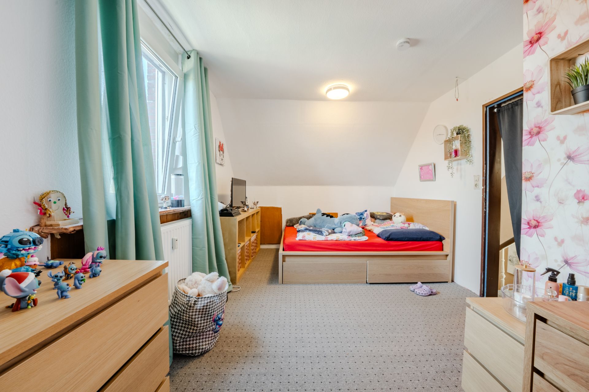 Kinderzimmer mit Bett, Spielzeug, Kommode und grünem Vorhang am Fenster.
