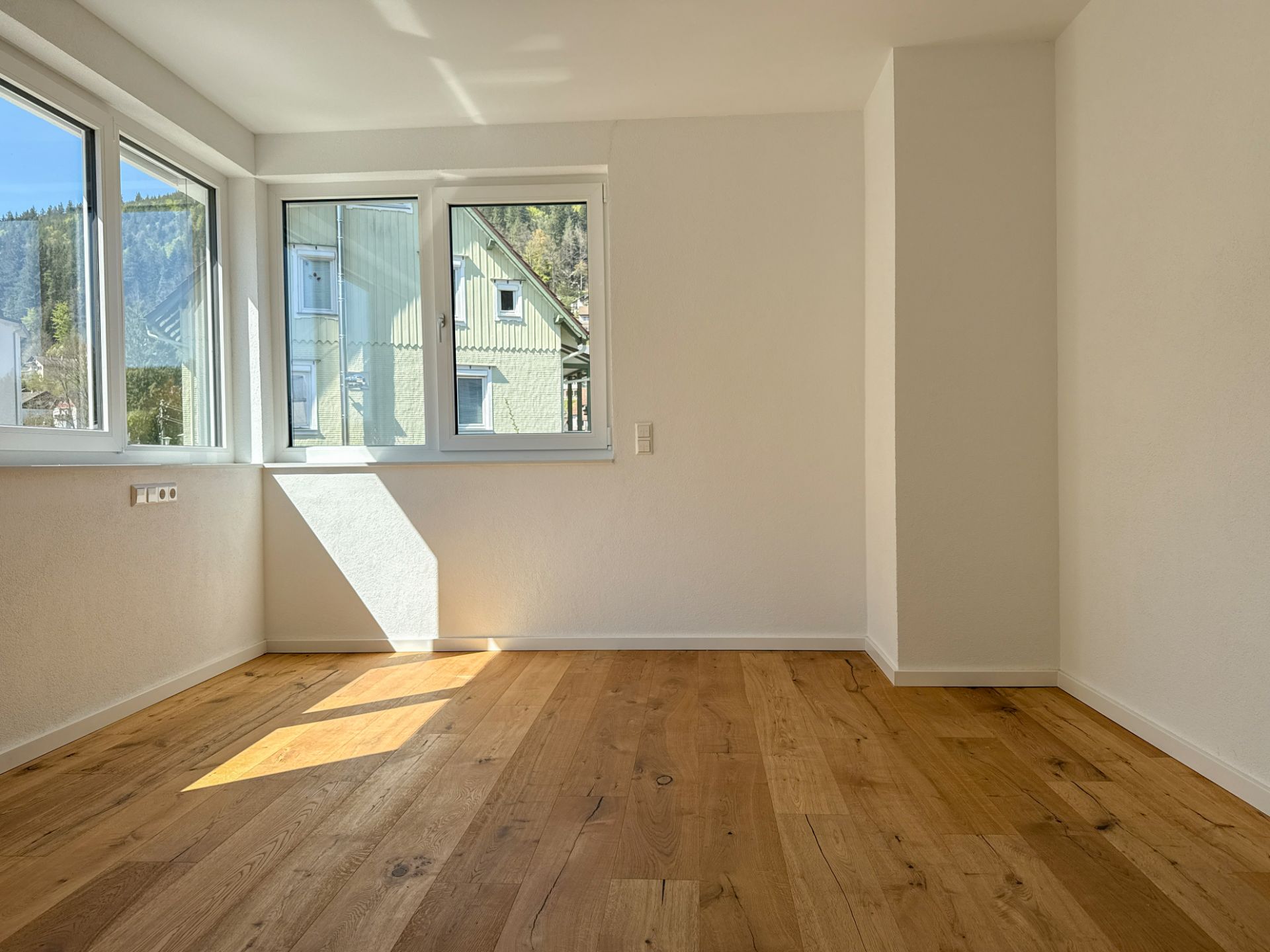Helles Zimmer mit großen Fenstern und Holzboden, Sonnenlicht fällt auf den Boden.