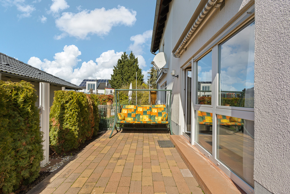Gepflasterte Terrasse mit Schaukelbank neben einem Haus, umgeben von grünen Büschen.