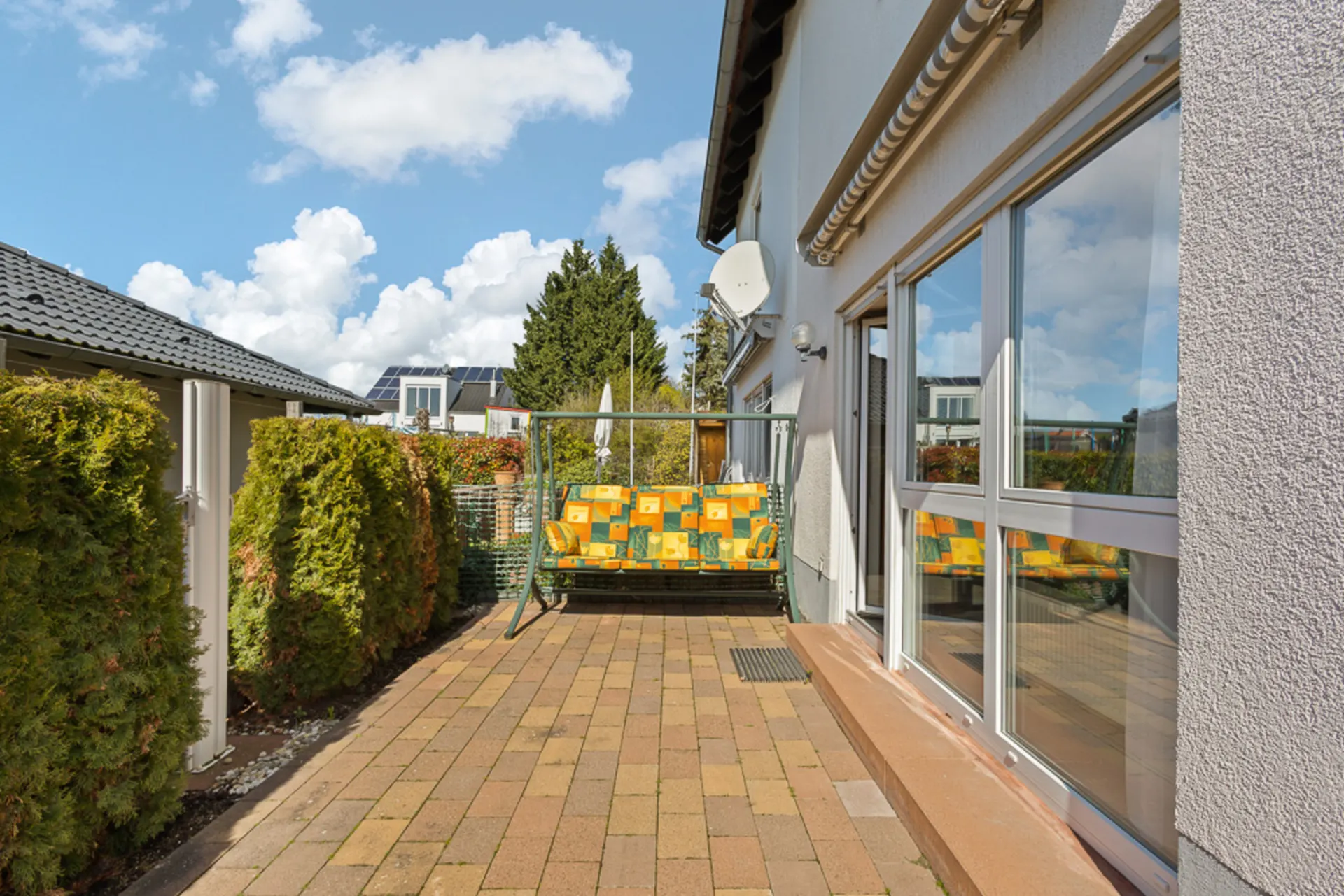 Gepflasterte Terrasse mit Schaukelbank neben einem Haus, umgeben von grünen Büschen.