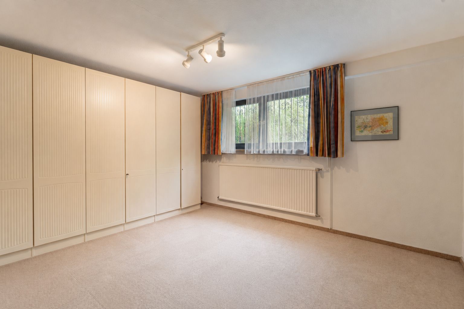 Helles Zimmer mit großem Einbauschrank, Fenster mit Gardinen und Bild an der Wand.