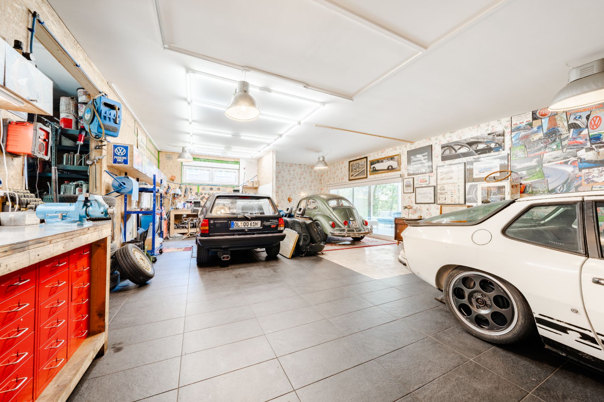 Große Garage mit drei Autos, Werkbank, Werkzeugen und vielen Autoplakaten an den Wänden.