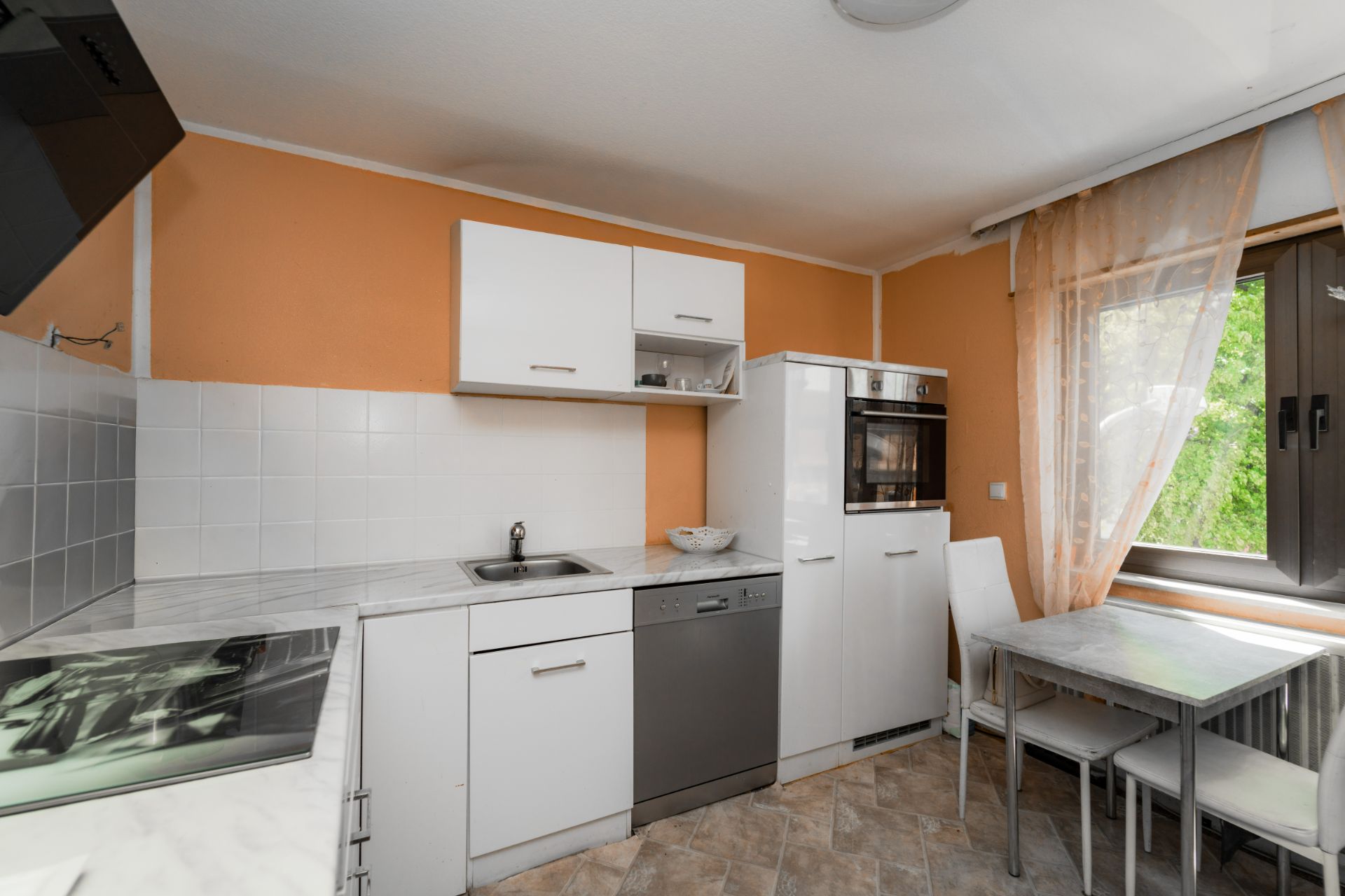 Helle Küche mit weißen Schränken, Spüle, Herd, Backofen und Esstisch am Fenster.