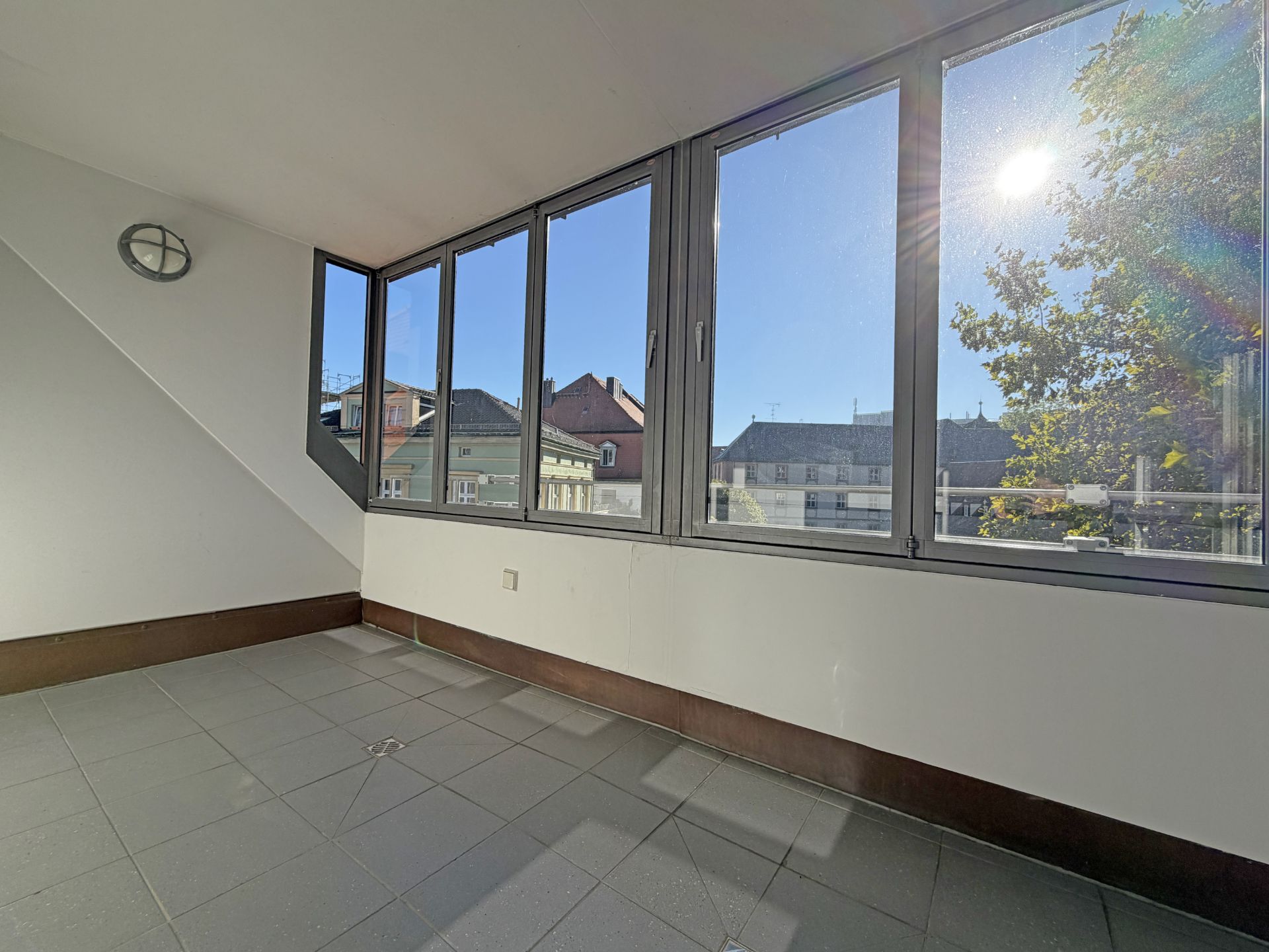 Heller, verglaster Balkon mit Blick auf Dächer und einen Baum bei Sonnenschein.