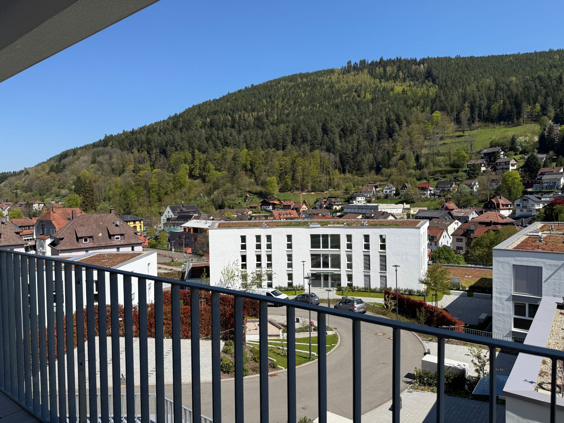 Blick von einem Balkon auf Wohnhäuser, Straße und bewaldeten Hügel im Hintergrund.