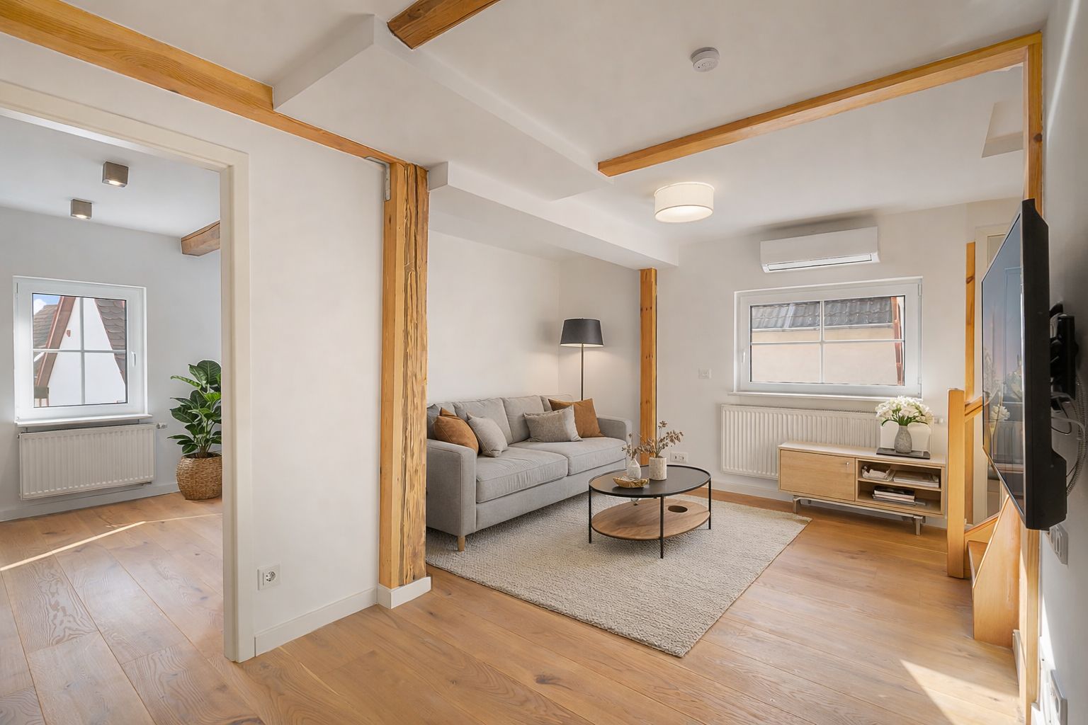 Helles Wohnzimmer mit grauem Sofa, Teppich, Couchtisch und großem Fenster, Holzboden und Deckenbalken.