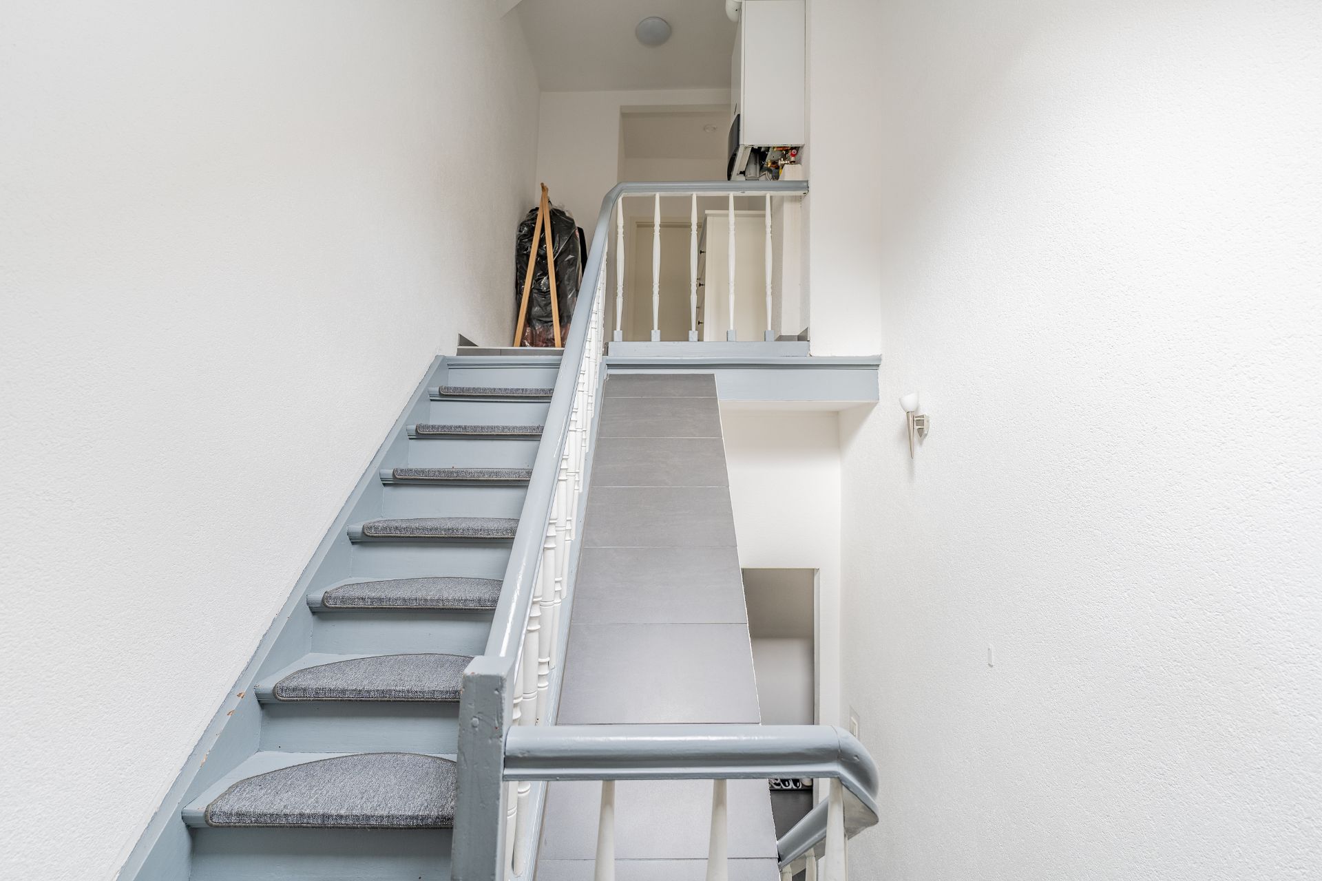 Graue Treppe mit Teppichstufen in einem hellen Flur, oben ein Geländer und einige Gegenstände.