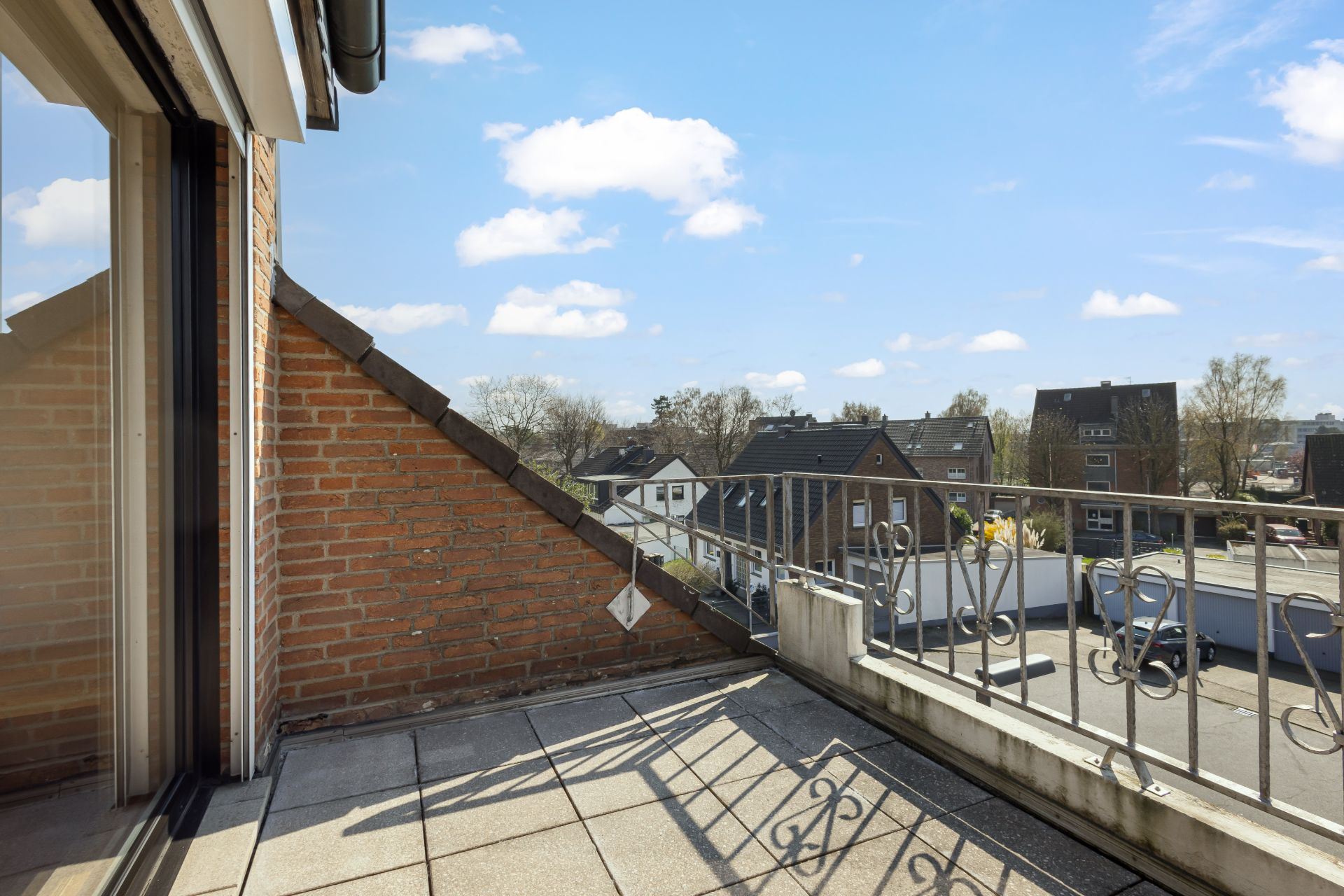 Kleiner Balkon mit Fliesenboden, Geländer und Blick auf Nachbarhäuser bei Sonnenschein.