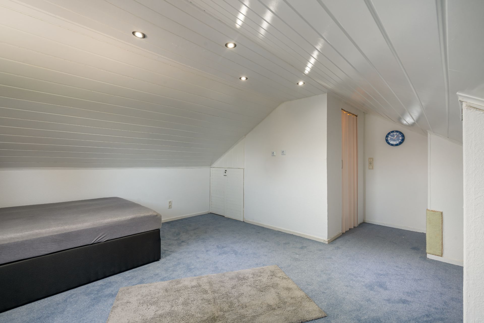 Helles Zimmer mit Dachschräge, grauem Teppich, Bett und Deckenleuchten.