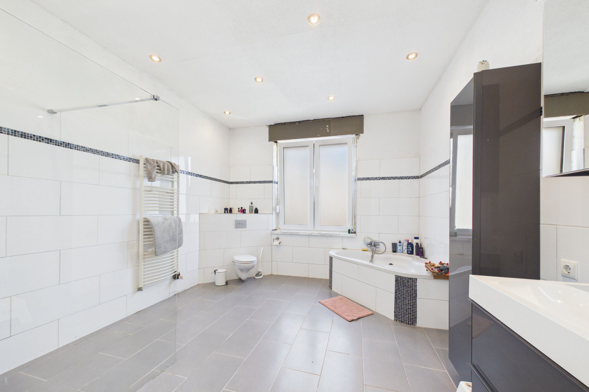 Modernes, weiß gefliestes Badezimmer mit Eckbadewanne, WC, Fenster und Handtuchheizkörper.
