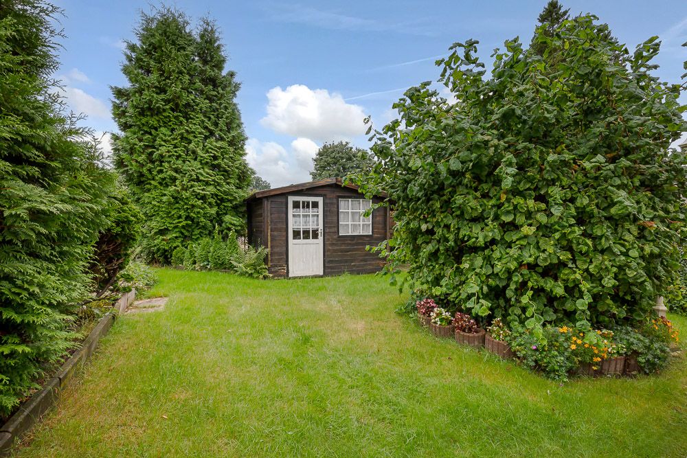Holzgartenhaus auf Rasen, umgeben von Büschen, Blumen und Bäumen.