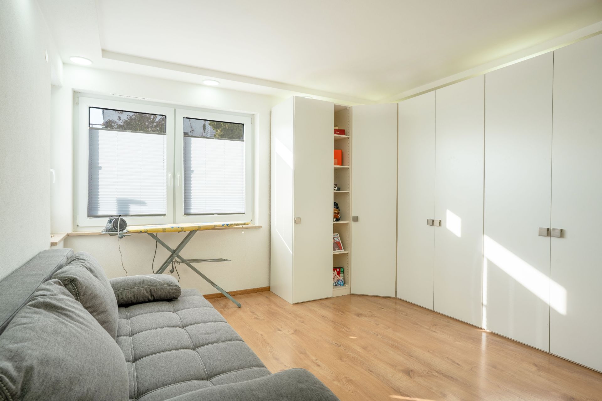 Helles Zimmer mit grauem Sofa, großem Schrank, Schreibtisch unter Fenstern und Holzfußboden.