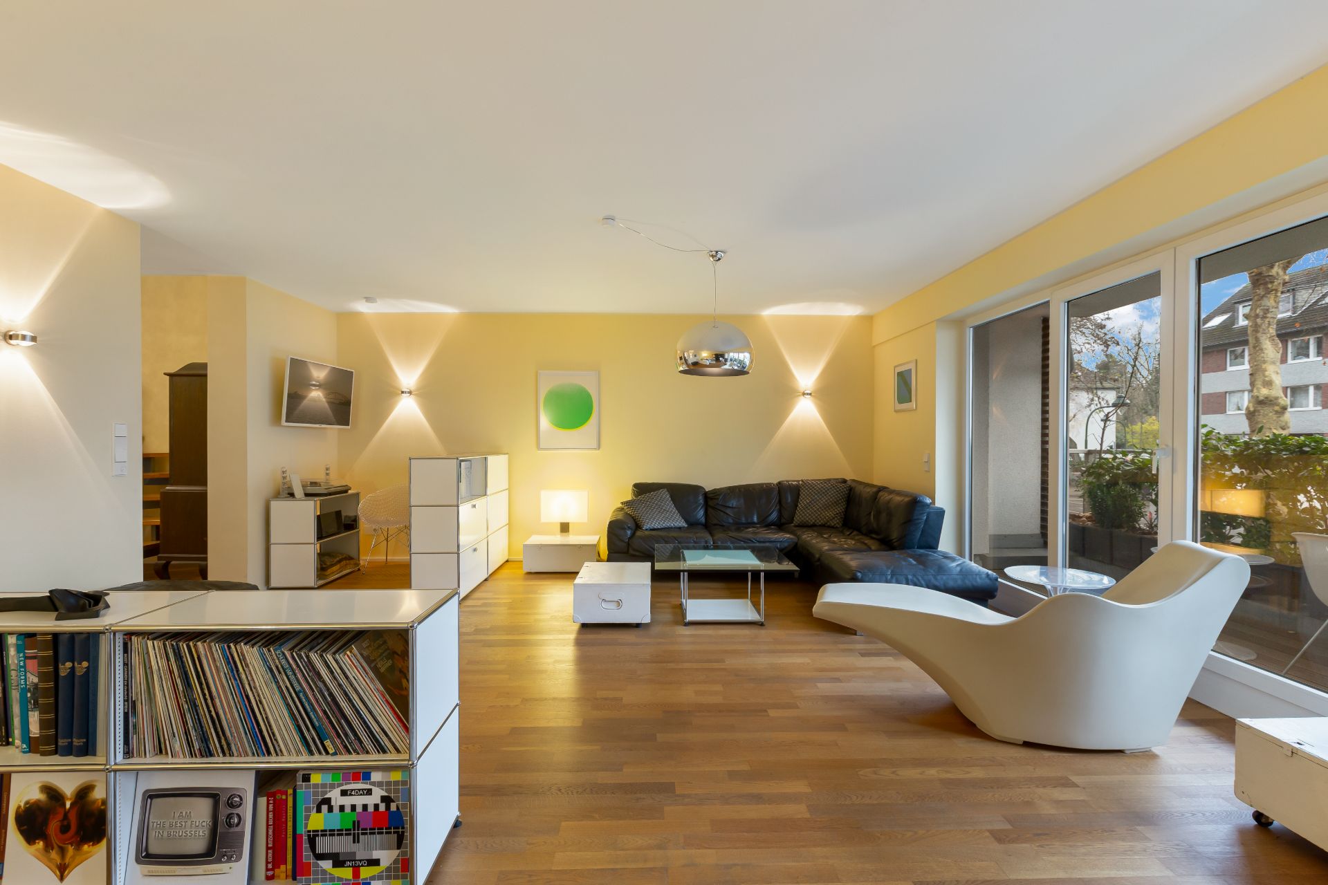 Modernes Wohnzimmer mit Holzboden, schwarzem Sofa, Sessel und Zugang zum Balkon.
