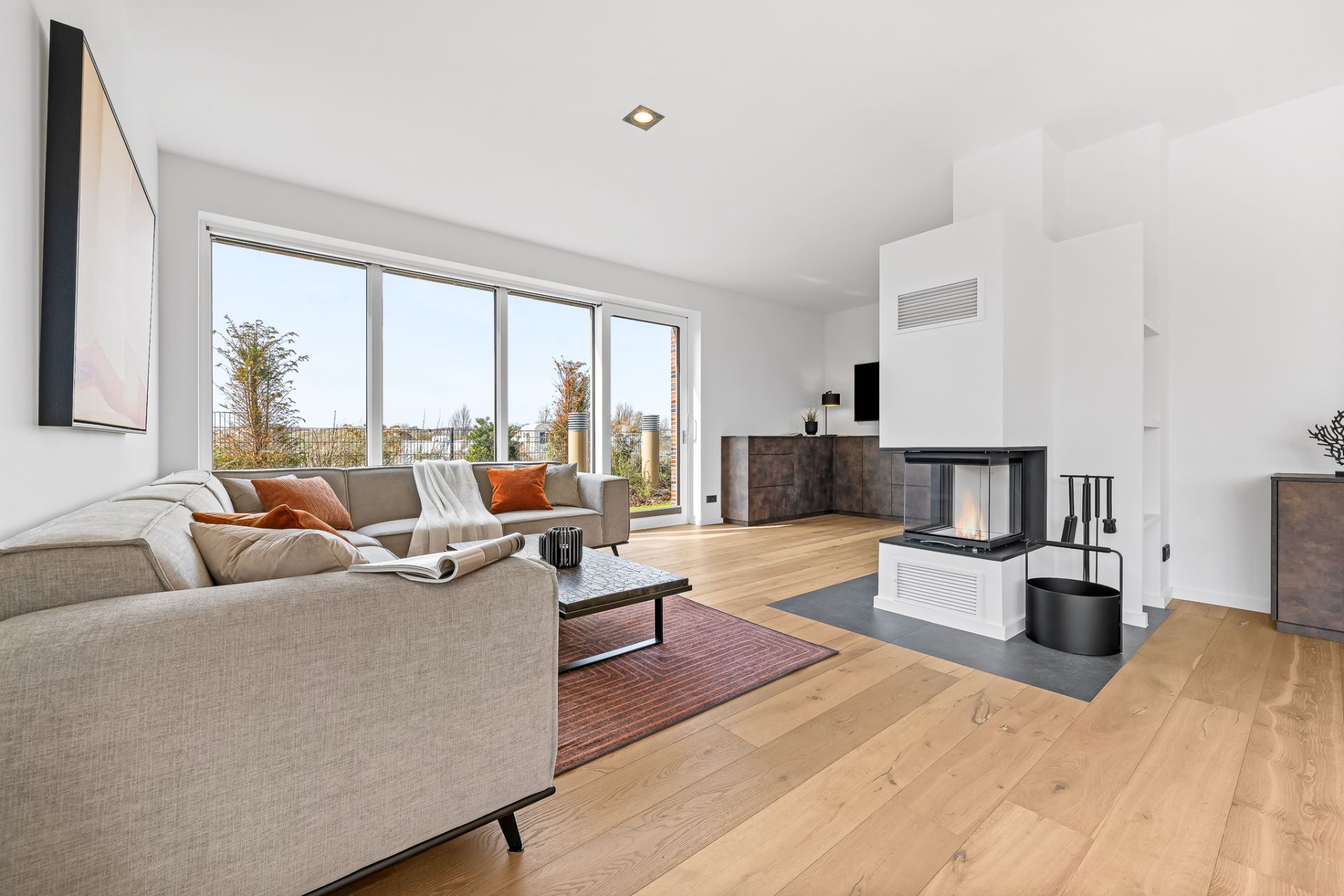 Helles Wohnzimmer mit Sofa, Kamin, Couchtisch und großen Fenstern mit Blick ins Freie.