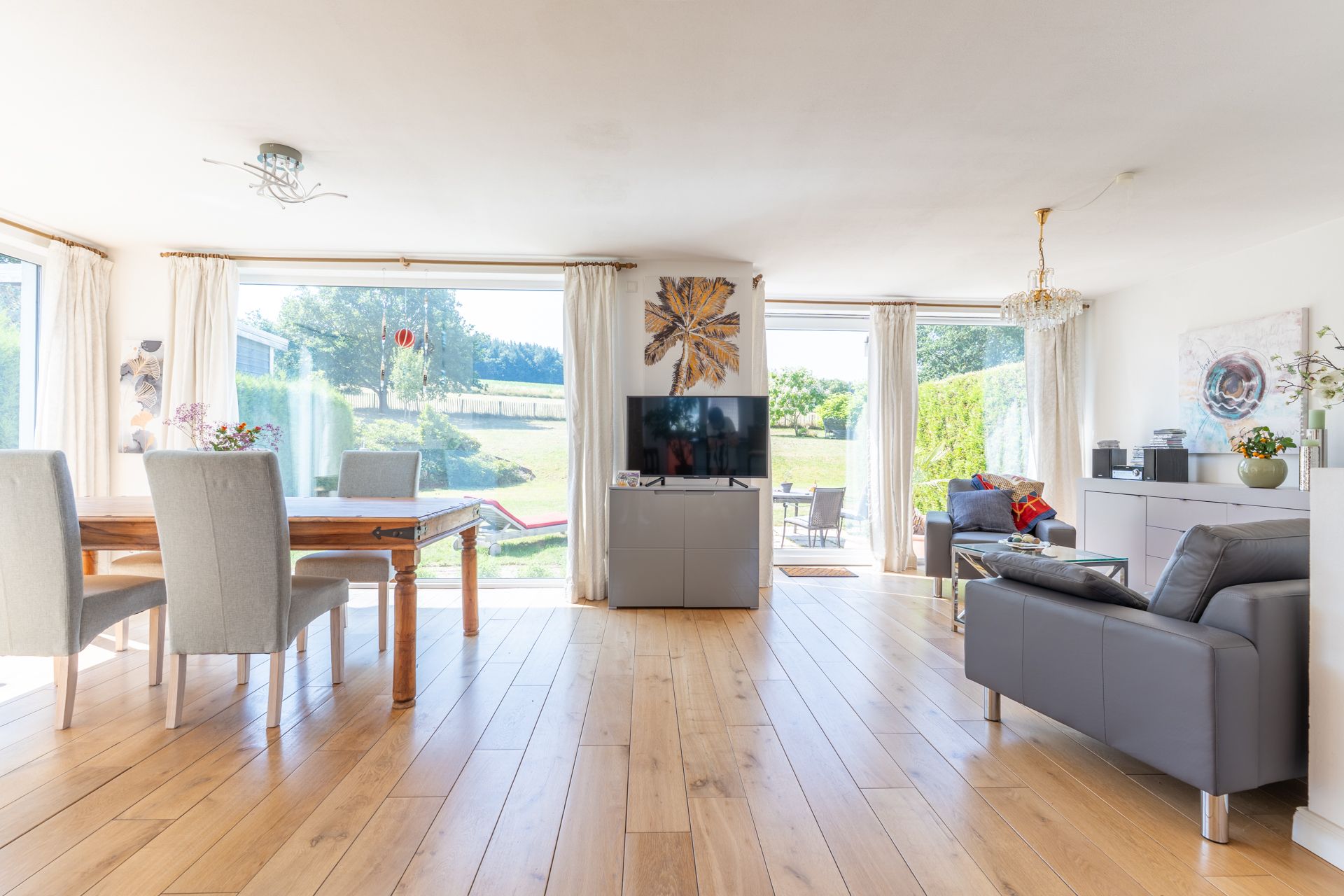 Helles Wohnzimmer mit Holzboden, grauen Sofas, Esstisch und großen Fenstern mit Blick ins Grüne.