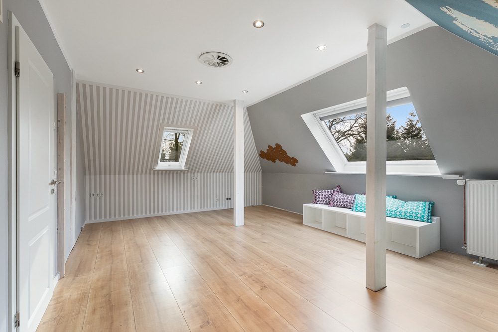 Helles Dachzimmer mit Holzfußboden, großen Fenstern und Sitzbank mit Kissen.