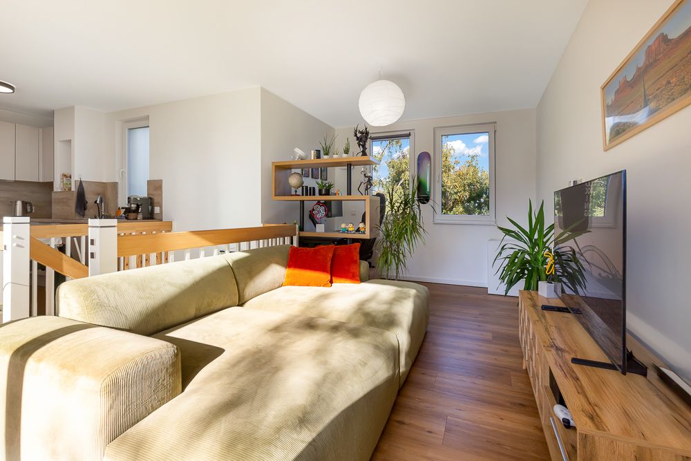 Helles Wohnzimmer mit großem Sofa, Fernseher, Pflanzen und Holzregalen, zwei Fenster im Hintergrund.
