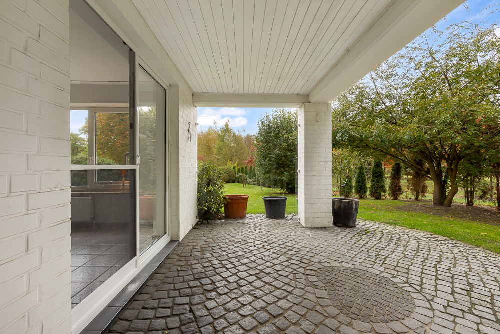 Überdachte Terrasse mit Pflastersteinen, mehreren Pflanzkübeln und Blick in einen Garten mit Bäumen.