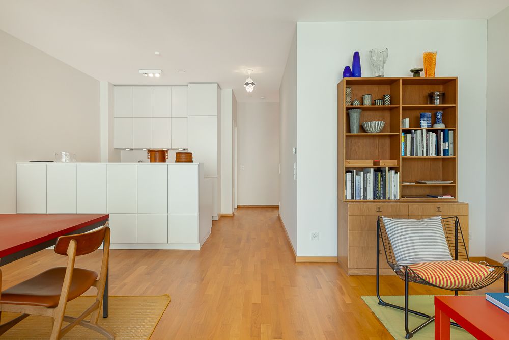 Modernes Wohnzimmer mit Bücherregal, Stuhl, rotem Tisch und offener Küche mit weißen Schränken.