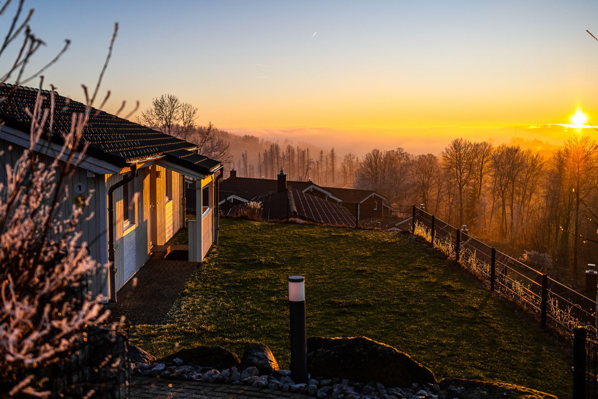 Haus mit Garten im Vordergrund, Sonnenaufgang und Bäume im Hintergrund.